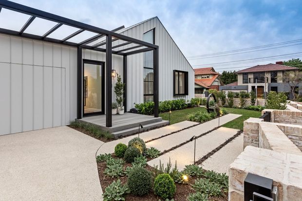 Contemporary residential exterior with a crisp white vertical-panel facade, steep gabled roof, black-framed glazing, and a covered outdoor entry terrace. The landscaped front garden features geometric pale paving paths, low succulents and clipped shrubs, while a light masonry boundary wall and neighbouring homes are visible beyond.