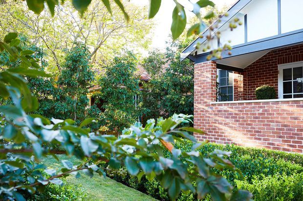 Exterior view of a brick residential building partially screened by lush garden foliage. The facade features red brick walls, dark-framed windows, a projecting covered corner or balcony-like element, and a contemporary roofline, with neatly clipped hedges and mature planting in the foreground.