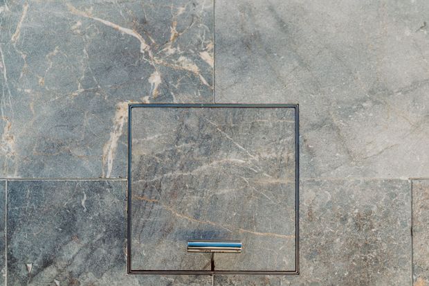 Close-up of a wall-mounted flush plate set into large-format grey-blue stone-look tiles, with subtle warm veining and a slim dark metal frame and actuator bar.
