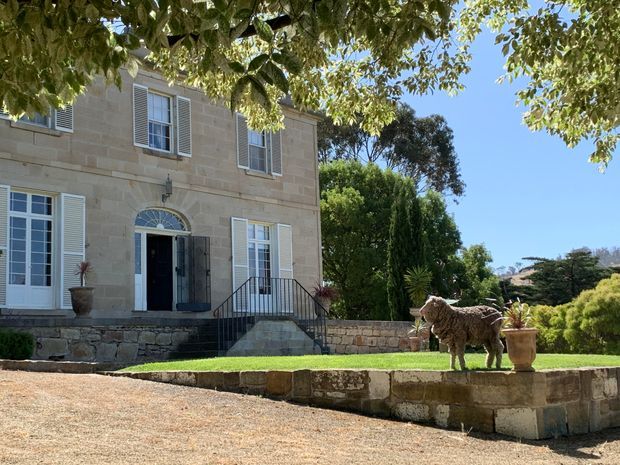 Exterior view of a substantial light-stone, two-storey residence with symmetrical window openings, white louvred shutters, and a classical arched entrance with fanlight. The home sits on a raised stone terrace with steps, low retaining walls, manicured lawn, mature garden planting, and a prominent sheep sculpture. Bright sunny setting with a formal, traditional character.