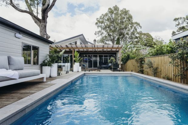 A contemporary backyard pool area viewed from the edge of a rectangular swimming pool. The space includes a timber deck with outdoor seating, a light-clad pavilion or house extension with extensive glazing, a timber pergola, potted tropical plants, and a timber privacy fence. Mature trees and a bright overcast sky surround the property.