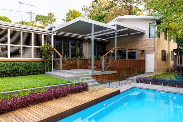 Exterior view of a contemporary brick house and landscaped backyard, featuring a covered outdoor entertaining deck, timber pool surround, and bright swimming pool. The home has broad louvred glazing, slim metal-framed patio structures, timber privacy screens, and layered garden planting.