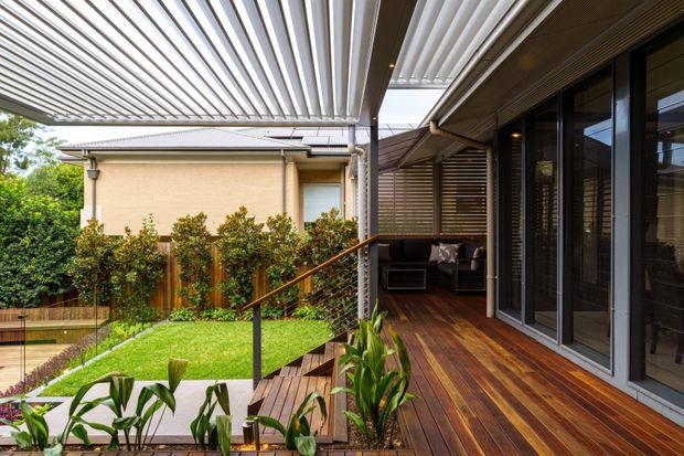 Contemporary covered outdoor living area with a timber deck, dark-framed sliding glass doors, integrated louvred pergola and privacy screens. The deck overlooks a landscaped lawn and planted garden, with steps and a slimline railing connecting the levels.