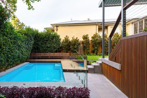 Landscaped residential courtyard with a bright blue swimming pool, timber pool decking and built-in bench seating, paved surrounds, planted screening, and a contemporary timber-and-metal privacy structure with stairs and glass balustrading. A traditional-looking house is visible beyond the garden.