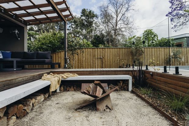 A landscaped outdoor entertaining area with a raised timber deck, dark outdoor seating, a timber pergola, and a sunken fire pit lined with pale gravel and concrete-edged bench seating. Timber privacy fencing and planting surround the compact courtyard-style space.
