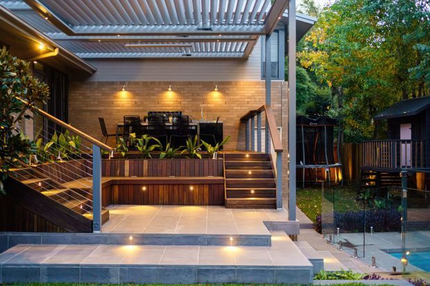 A landscaped residential outdoor entertaining area with a raised timber-deck dining zone, integrated planter boxes, illuminated stairs, tiled paving, and a louvred pergola roof. The space opens toward a fenced pool area and is surrounded by mature planting. Warm wall and step lighting highlights the timber and exposed brick finishes.