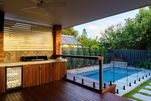 Covered outdoor entertaining area with a built-in timber outdoor kitchen and bar, overlooking a compact swimming pool. The space features dark timber decking, warm timber cabinetry, brick columns, a patterned tiled splashback, glass pool fencing, and a slatted privacy screen. A landscaped suburban garden and neighbouring roof are visible beyond the pool.