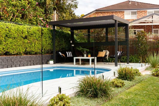 A landscaped backyard with a bright blue swimming pool and a contemporary covered outdoor lounge pavilion. The pavilion has a slim dark metal frame, flat roof, horizontal privacy screens, and casual seating around a small white table. Hedging, ornamental grasses, paved pool surrounds, and a neighbouring brick house are visible.