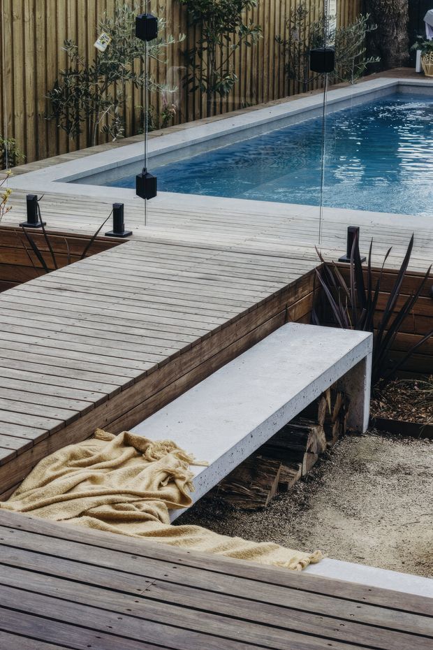 A landscaped outdoor pool area with a rectangular swimming pool bordered by pale concrete coping and extensive timber decking. A weathered timber privacy fence, slender planted trees, dark pendant-style outdoor lights, and a simple concrete bench create a contemporary natural setting. A folded beige throw rests on the bench and decking, adding a relaxed poolside character.