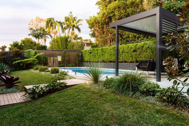 A landscaped residential backyard featuring a rectangular swimming pool, timber decking and pathways, manicured lawn, layered tropical and subtropical planting, a timber privacy fence, and a contemporary dark-framed covered outdoor pavilion with translucent roofing and integrated screening. The setting is lush, private, and resort-like.