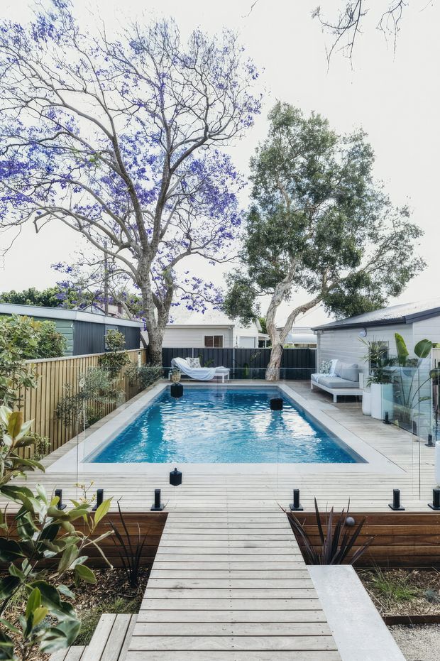 A landscaped backyard centred on a rectangular swimming pool, viewed along a light timber boardwalk. The pool is surrounded by pale decking, dark timber raised garden beds, potted plants, outdoor lounge seating, privacy fencing and low modern single-storey structures. Mature trees provide a leafy setting, including a prominent flowering tree overhanging the pool.