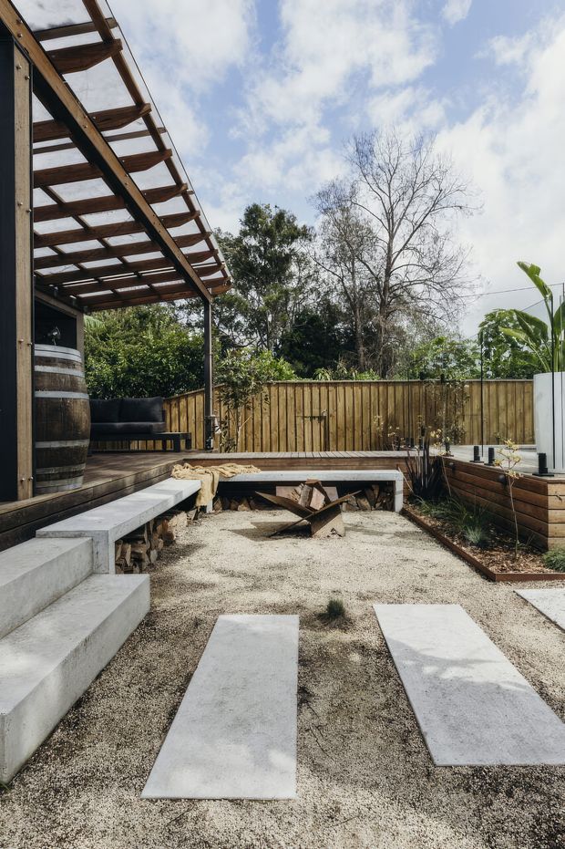 A landscaped residential courtyard or backyard with a timber pergola and deck, raised concrete seating steps, a built-in timber bench, gravel ground cover with large rectangular stepping slabs, timber fencing, planted garden beds, and an outdoor fire pit area. The space has a warm, rustic contemporary outdoor entertaining character.