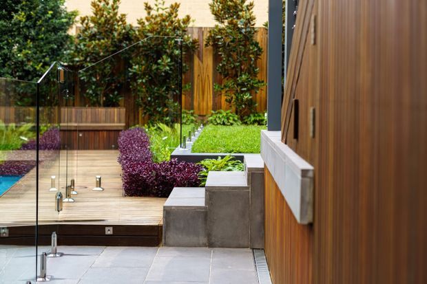 A contemporary landscaped outdoor courtyard or pool terrace viewed from beside a timber-clad building. Large-format grey paving leads to a timber deck and a partially visible blue pool, enclosed by frameless glass balustrades. Tiered concrete planters, lush green planting, purple ornamental shrubs, and warm vertical timber screening create a refined, private garden setting.