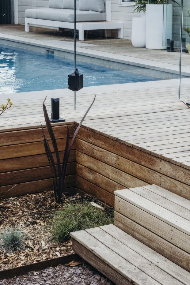 A landscaped outdoor poolside area with a light timber deck, integrated timber planter and stepped bench seating. A frameless glass pool barrier separates the deck from a blue swimming pool, with a minimalist white outdoor sofa and planters beyond.