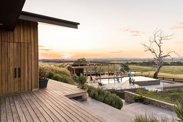 A contemporary rural property viewed from a timber-clad deck, with a broad landscape outlook, expansive paved terrace, swimming pool, covered outdoor entertaining area, and a simple dark roofline. The scene is photographed at sunset and combines warm timber surfaces with muted natural colours.