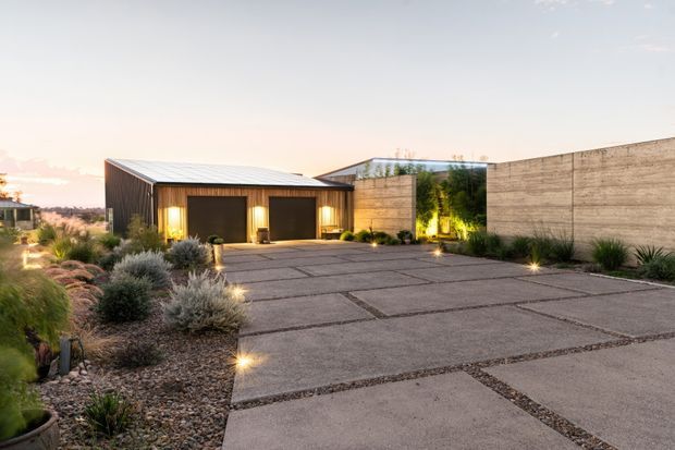 A contemporary single-storey property viewed from a broad paved driveway or forecourt at dusk. The building combines a dark metal-clad garage volume with warm timber entry detailing, a low-slung metal roof, and large textured concrete walls enclosing a landscaped courtyard. Gravel beds, sculptural grasses, shrubs, and subtle ground lighting soften the hardscape.