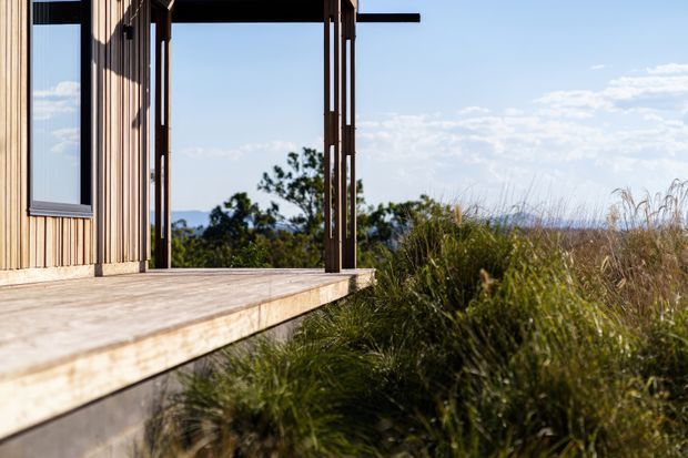Exterior view of a contemporary timber-clad structure with a broad timber deck projecting into native grasses. Slim dark-framed glazing and an open corner frame distant hills and vegetation, creating a strong indoor–outdoor connection. The image is photographed from outside and focuses on the building envelope, deck, and surrounding landscape.