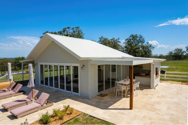Bright exterior view of a single-storey white weatherboard home with a pitched metal roof, extensive glazed doors, and a covered outdoor entertaining area opening onto a paved terrace. Sun loungers and a pool edge are visible in the foreground, with landscaped grounds and open countryside beyond.