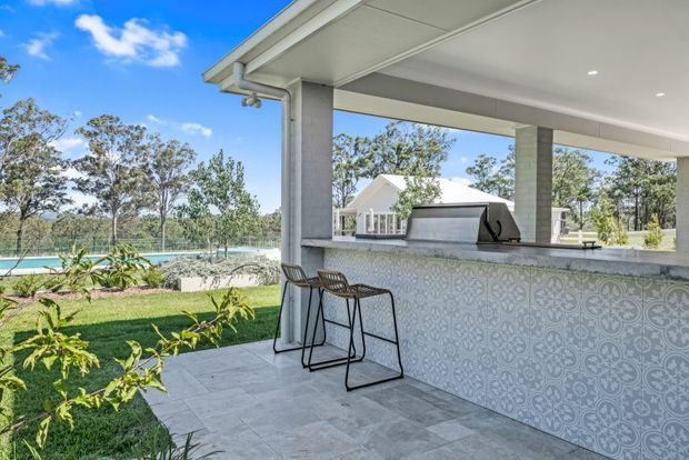 A bright covered outdoor entertaining area with a built-in tiled bar, pale stone-look benchtop, barbecue, and two black metal bar stools. The space is supported by white-painted masonry columns and has a light grey tiled floor and ceiling downlights. A swimming pool and landscaped garden are visible beyond the open sides, with a detached light-coloured gabled structure in the background.