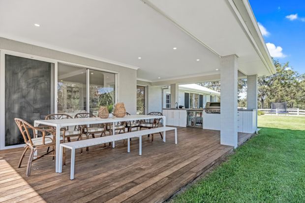 A bright covered outdoor entertaining area with a timber deck, long dining table and woven chairs, opening to a built-in outdoor kitchen and grassy yard. The space has a white rendered structure, white-framed glazing, recessed ceiling lights and a broad low-pitched roof.