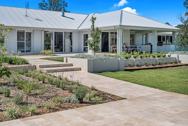 Single-storey light-coloured residential home with a broad corrugated metal hipped roof and covered verandah. The facade has extensive glazed doors and windows, landscaped raised garden beds, paved paths and steps, and a manicured lawn under bright blue skies.
