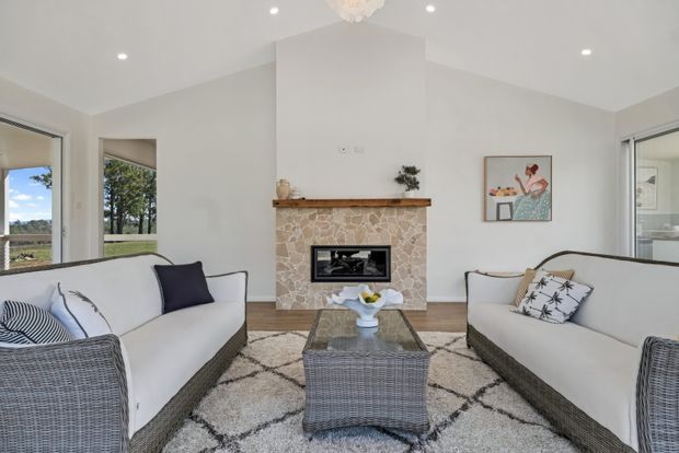 Bright, spacious living room with two matching white upholstered sofas featuring woven rattan-style bases, arranged around a glass-topped woven coffee table. A stone-clad fireplace with timber mantel forms the focal point, with a large geometric rug, recessed ceiling lights, neutral walls, and expansive windows overlooking greenery. Coastal-inspired artwork and cushions add understated colour.