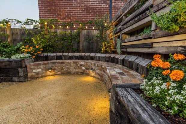 A landscaped courtyard garden with a curved, built-in brick seating wall enclosing a compact gravel area. Raised timber planter beds, planted greenery and orange flowers line the perimeter, while warm string lights and integrated wall lighting create an inviting evening atmosphere. A red-brick building wall and rustic horizontal timber screening form the backdrop.
