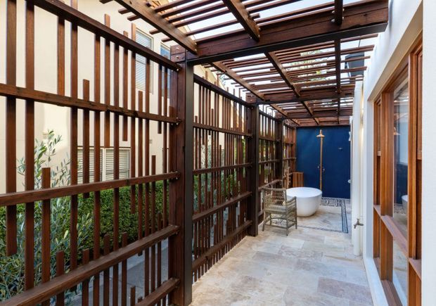 A narrow, partially enclosed outdoor passage or courtyard with pale stone paving, dark timber privacy screens, and a timber pergola overhead. The space includes a freestanding white bathtub at the far end, a small woven chair, lush planting behind the screens, and blue and timber-framed doors along the building. The design creates a sheltered, private outdoor bathing area with strong linear timber detailing.
