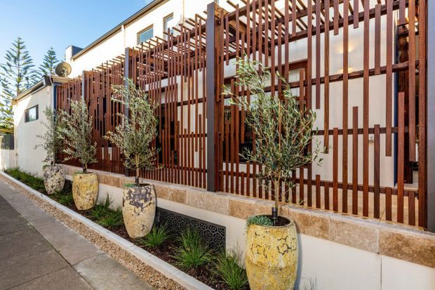 Exterior view of a contemporary multi-level residence with a prominent timber slatted privacy screen and pergola-like facade structure. White rendered walls, dark window frames, stone-look base detailing, and large textured planter pots with small trees line the narrow landscaped frontage beside a paved walkway.