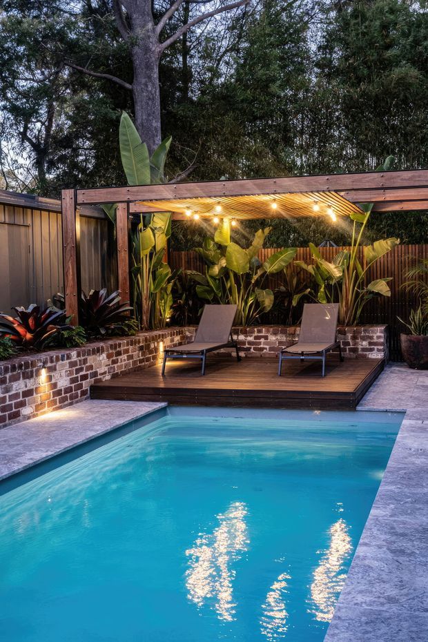A landscaped backyard pool area at dusk, featuring a rectangular turquoise swimming pool edged with pale paving, a raised dark timber deck with two lounge chairs, and a timber pergola fitted with warm string lighting. Brick retaining walls, timber fencing, and dense tropical planting create a private resort-like outdoor setting.
