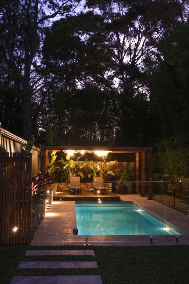 Twilight view of a landscaped residential backyard with a rectangular swimming pool enclosed by frameless glass fencing. The pool terrace is paved in large rectangular tiles and approached by stepping stones through lawn. A timber-framed, warmly lit cabana or outdoor entertaining structure sits behind the pool, surrounded by tropical planting, brick retaining walls and dense mature trees. The scene has a dark, atmospheric palette with warm architectural and landscape lighting.