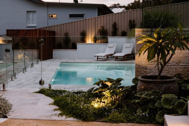 A landscaped residential courtyard with a rectangular swimming pool, pale stone paving, rendered privacy walls and timber fencing. Two loungers sit beside the pool, while layered tropical planting and subtle garden lighting create an intimate evening outdoor setting. A residence is visible beyond the courtyard.