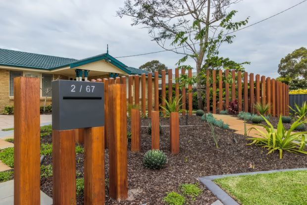 Street-facing residential property entrance with a dark grey mailbox marked 2 / 67, surrounded by a contemporary vertical timber slat fence. A single-storey brick home with a teal pitched roof is visible behind the landscaped front garden, which includes mulch beds, paving, grasses, succulents, and agave-like plants.
