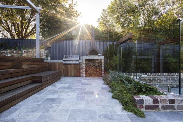 A landscaped outdoor entertaining courtyard with broad pale stone paving, tiered dark timber seating, a built-in wood-fired fireplace, dark metal fencing, brick and stone retaining walls, planting, and a glass pool or garden enclosure. The space is sunlit and framed by mature trees.