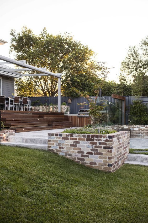 A landscaped residential backyard with a stepped outdoor entertaining area. The space includes a timber deck and stairs, outdoor dining furniture beneath a slim pergola or shade structure, dark metal fencing, a built-in barbecue area, glass balustrading, paved terraces, and raised garden beds constructed from multicoloured recycled-style brick. A neat lawn occupies the foreground, with established trees and planting around the perimeter.