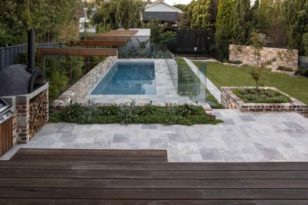 A landscaped residential backyard viewed from an elevated timber deck. The garden features a rectangular swimming pool surrounded by pale grey stone paving, frameless glass pool fencing, raised brick and timber garden beds, mature greenery, and a built-in outdoor pizza oven or fireplace with a stainless-steel barbecue. Dark timber decking and black fencing create a refined contemporary outdoor entertaining setting.