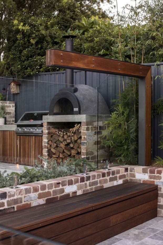 A landscaped outdoor entertaining area with a built-in wood-fired pizza oven, stainless-steel barbecue, timber cabinetry, and a dark-framed outdoor kitchen structure. A timber-clad overhead beam and posts support a black flue, while a low exposed brick wall doubles as a raised planter and boundary around a dark timber deck or bench. Dense planting and charcoal corrugated fencing create a private, warm, rustic-modern setting.