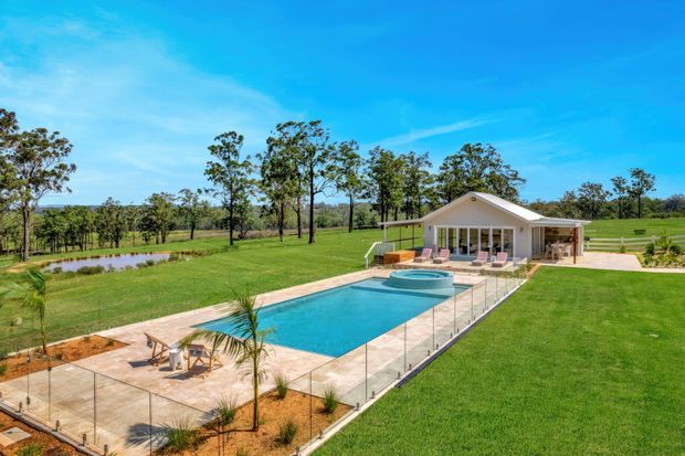 A bright aerial view of a landscaped rural property centred on a rectangular swimming pool with an attached circular spa, pale paved pool terrace, glass safety fencing, sun loungers, and a small single-storey pool pavilion. Expansive manicured lawns, young palms, scattered trees, a pond, and open fields surround the outdoor recreation area beneath a clear blue sky.