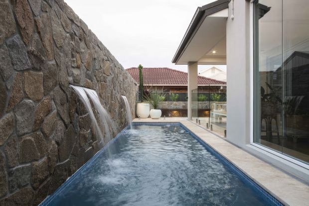 A narrow courtyard pool runs alongside a contemporary glass-walled building, with multiple water features cascading from a tall rough-stone masonry wall. The space includes pale tiled paving, white rendered walls, large sliding glass doors, and a tiled pitched-roof building visible beyond the courtyard.