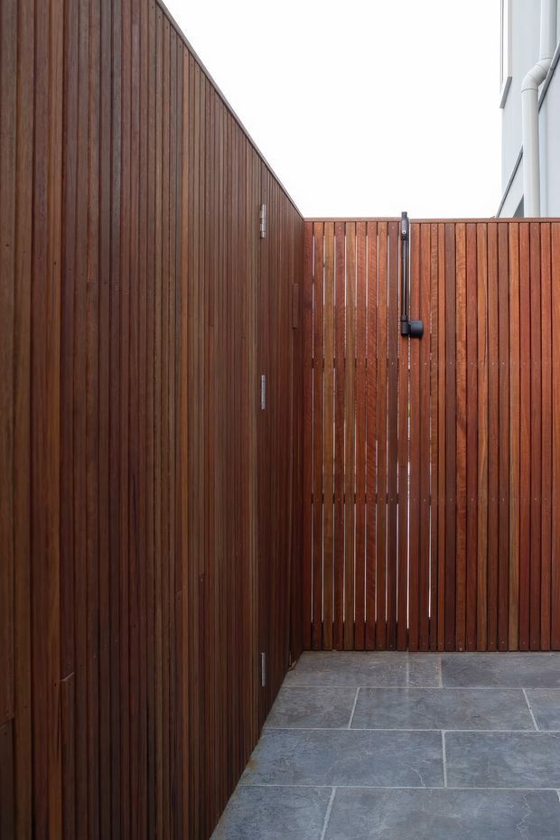 A narrow enclosed outdoor passage or courtyard with tall vertical timber privacy fencing and a matching timber gate. The fencing has a rich reddish-brown stained finish, while the ground is paved with large grey stone tiles. A small black gate latch and hardware are visible, with a partial light-coloured building wall at the right edge. The composition is minimalist and architectural, focused on the timber screening and paving.
