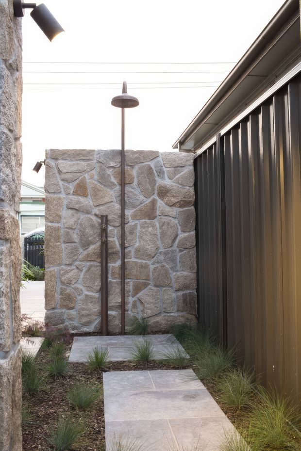 A narrow landscaped outdoor passage with a freestanding stone privacy wall, outdoor shower, concrete stepping slabs, ornamental grasses, and dark vertical metal cladding along the adjacent building. Wall-mounted and overhead lighting create a refined contemporary courtyard-like setting.
