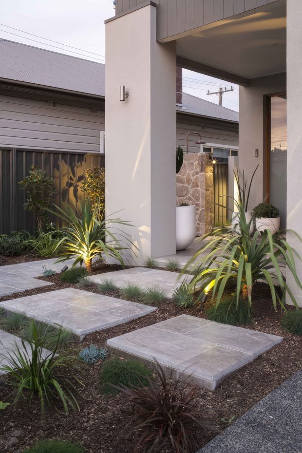 A contemporary residential entry courtyard with large pale stone stepping slabs set through a landscaped garden bed. Tall white rendered columns support a covered porch, with dark vertical metal fencing, timber-framed glazing, a stone feature wall, exterior lighting, and drought-tolerant ornamental planting. The image is viewed from the outdoor approach toward the house entrance.