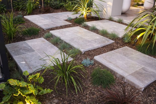 Contemporary landscaped garden path featuring large rectangular pale stone pavers set among dark mulch, ornamental grasses, succulents, and tropical-style planting. White rendered architectural elements are visible in the background.