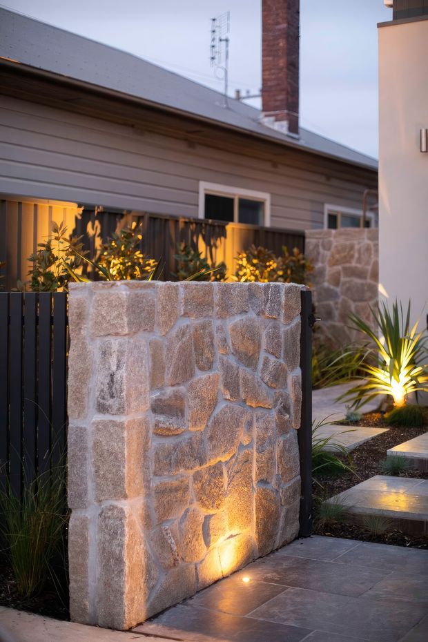 A landscaped residential entry courtyard at dusk, featuring a low rough-cut natural stone wall with integrated warm uplighting, dark vertical metal fencing, grey paving, architectural planting, and concrete stepping slabs. A grey weatherboard house with a pitched roof and brick chimney is visible behind, alongside a contemporary rendered building element.