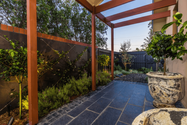 A landscaped outdoor courtyard or side garden with a dark stone-paved walkway, timber pergola, raised dark fencing, and dense planting. Large weathered stone urns sit beside the path, while warm sunlight highlights the timber structure and garden edges.
