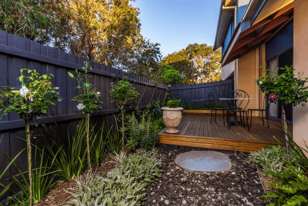 A compact landscaped courtyard beside a modern home, featuring a raised timber deck with a small metal café table and chairs, a large urn planter, dark timber privacy fencing, layered planting, and a circular concrete stepping pad set in a pebble garden. The house has rendered walls, dark-framed glazing, and exposed timber eaves.