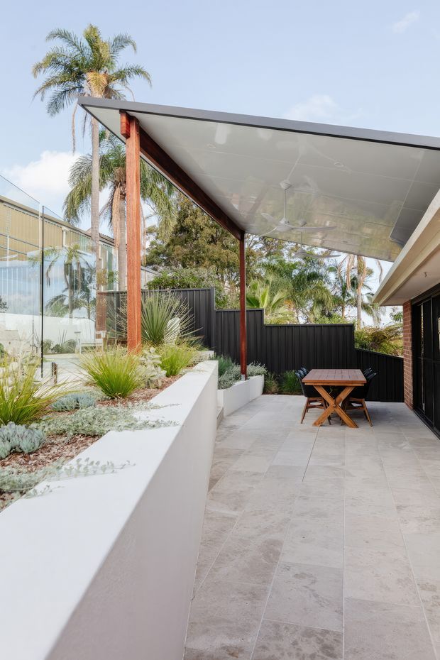 A bright landscaped outdoor courtyard or patio beside a modern home. Large pale stone-look paving extends beneath a broad flat-roofed canopy supported by slender timber posts, with a ceiling fan overhead. Raised white rendered planter beds contain drought-tolerant grasses and succulents, while black vertical-panel fencing provides privacy. A small timber folding table and chairs sit near dark-framed glazed doors.