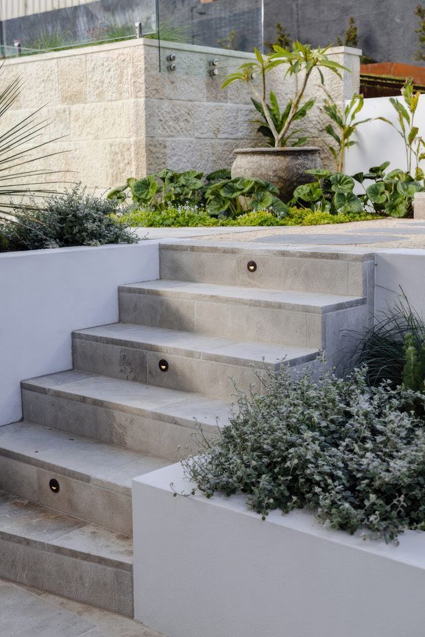 A landscaped contemporary outdoor stairway with broad light-grey tiled steps, integrated circular step lights, white rendered planter walls, and lush layered planting. At the upper landing, a textured light-stone wall incorporates outdoor shower fixtures behind a glass screen, with a large ceramic planter and tropical greenery. The space has a clean, refined courtyard-garden character.