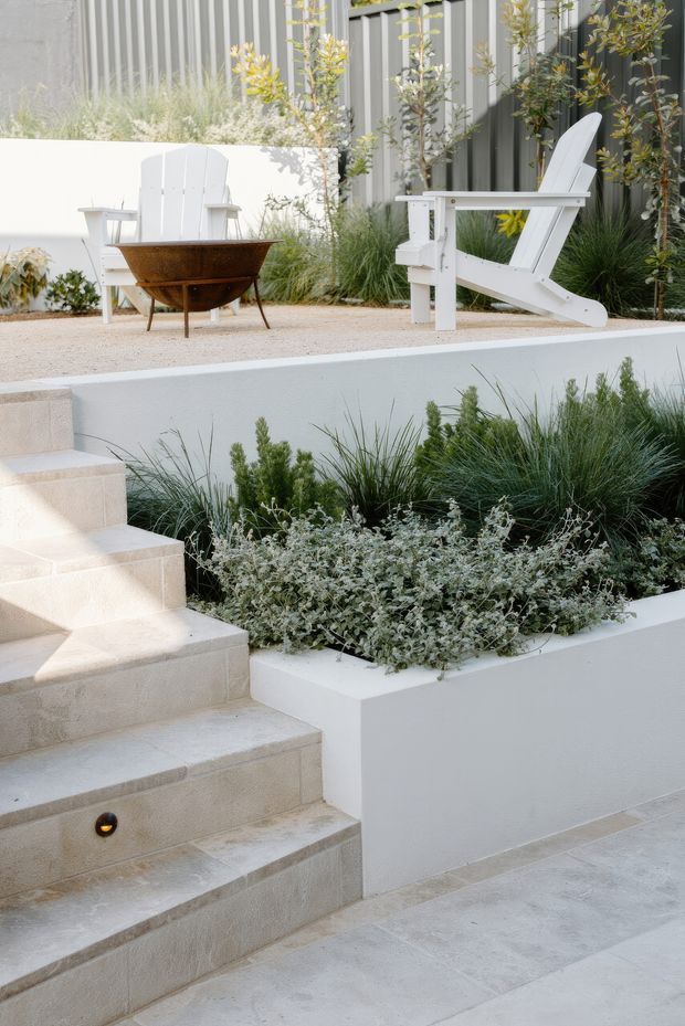 A contemporary landscaped outdoor terrace with broad pale stone steps, raised white rendered planters, ornamental grasses and silvery groundcover. At the upper level, white Adirondack-style chairs surround a rusted steel bowl fire pit, backed by dark vertical metal cladding and a light rendered wall.