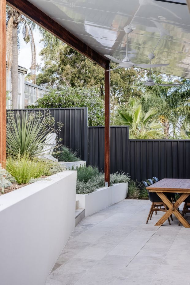 A landscaped outdoor dining terrace beneath a covered pavilion, featuring pale large-format tiled paving, white rendered raised garden beds, dark corrugated metal privacy fencing, timber posts and beams, and multiple ceiling fans. Drought-tolerant planting with palms and ornamental grasses surrounds a timber outdoor dining table and chairs.